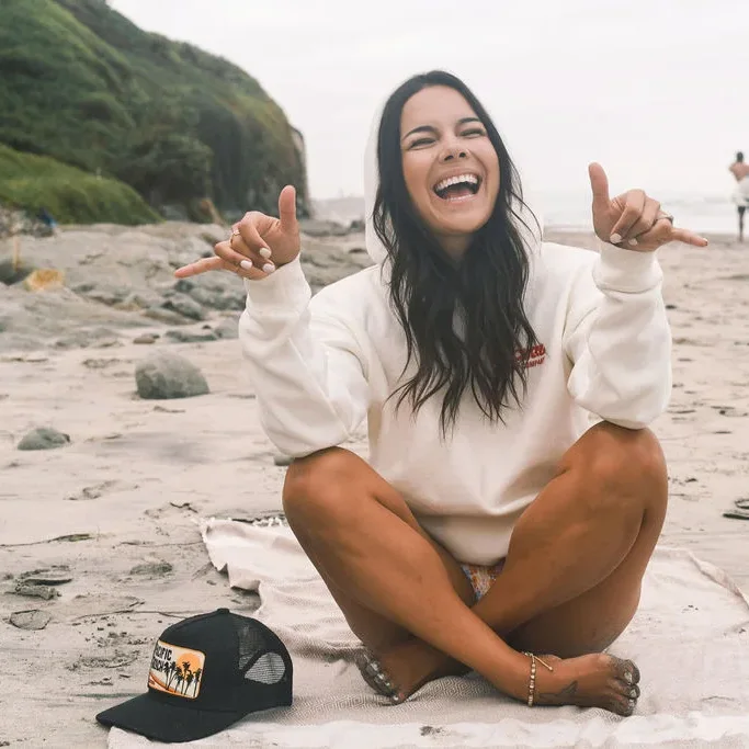 Founder, Jessica is playfully sitting on the beach with a big smile in a oversized hoodie